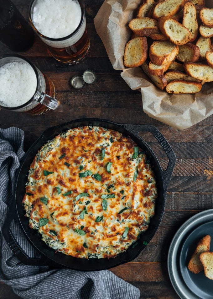 6 cheese spinach artichoke dip in a cast iron pan with a bowl of crostini