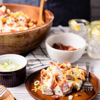 loaded potato salad served on a wooden plate, in front of a large wooden serving bowl