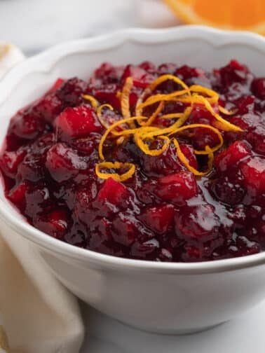 apple orange cranberry sauce in a white bowl next to a yellow and white striped towel. Orange wedges and a bottle of orange liqueur are in the background.