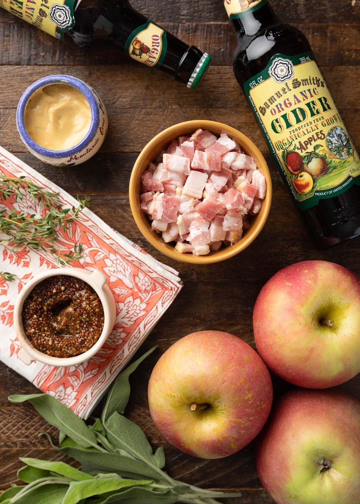 overhead viewe of bottles of hard cider, crocks of Dijon and grainy mustard, chopped bacon, apples, and herbs on a wood board