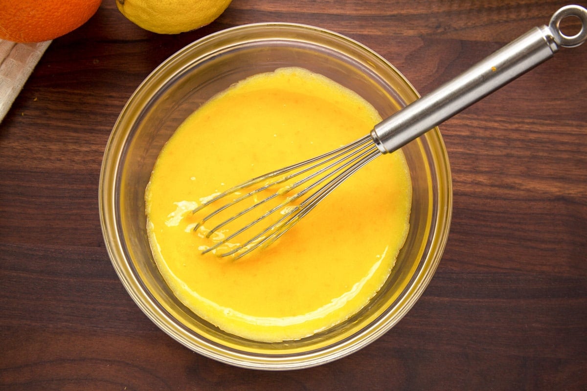 orange curd in a glass bowl with a stainless steel whisk on a dark wood cutting board