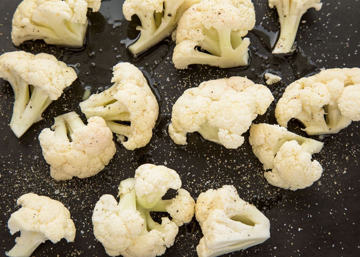 raw cauliflower florets on a black baking sheet with olive oil, salt, and pepper