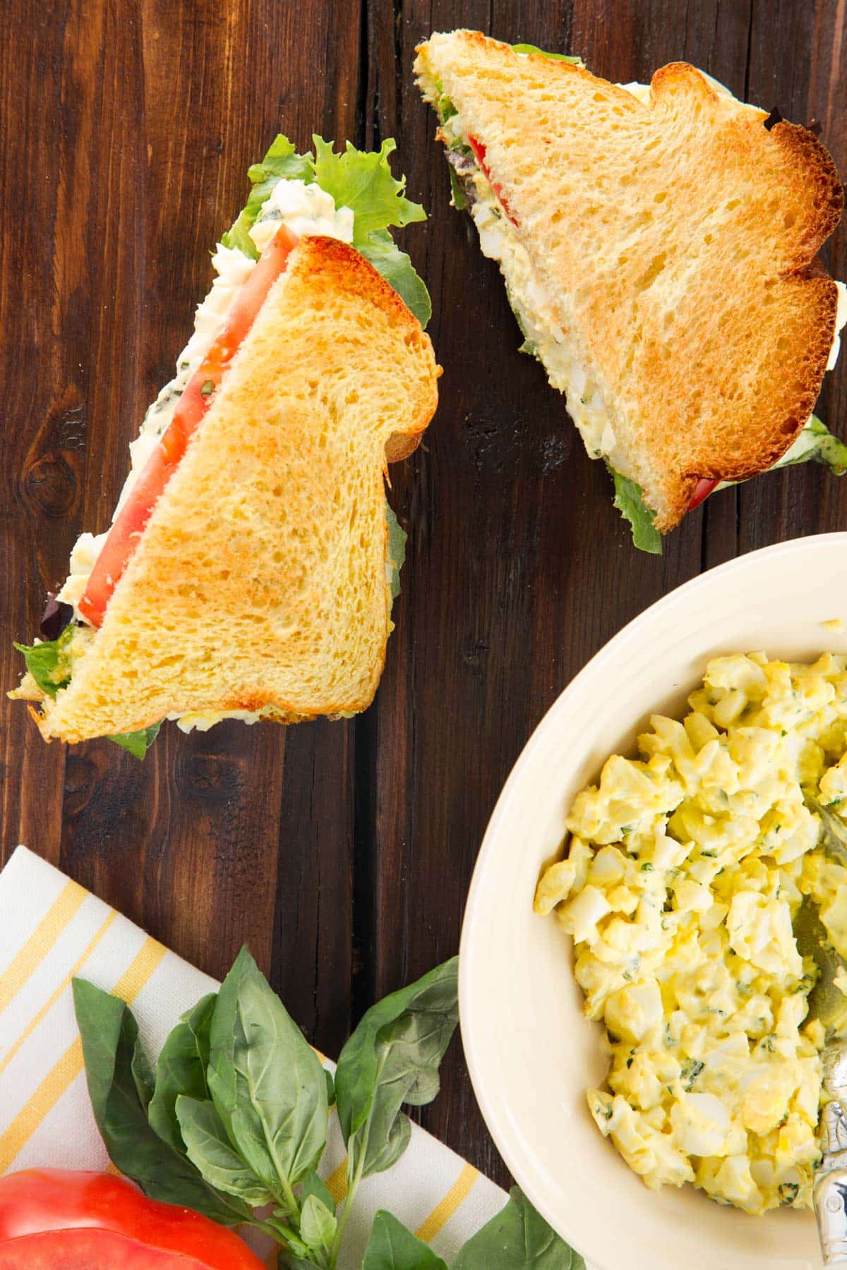 two halves of a basil egg salad sandwich on brioche toast on a wood board next to a bowl of egg salad 