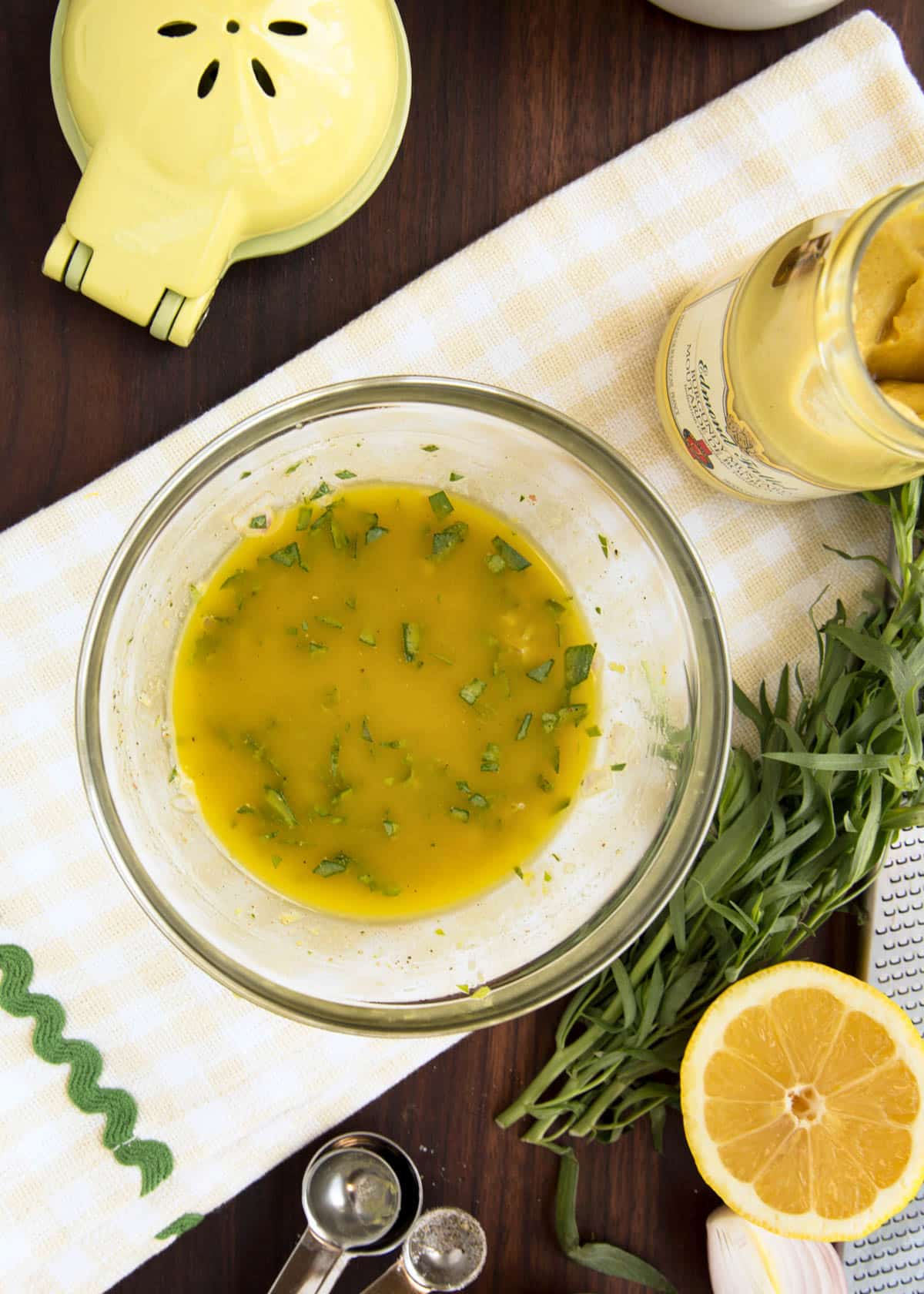 overhead photo of tarragon vinaigrette in a bowl on an ivory checked napkin surrounded by lemons, a jar of mustard, and herbs