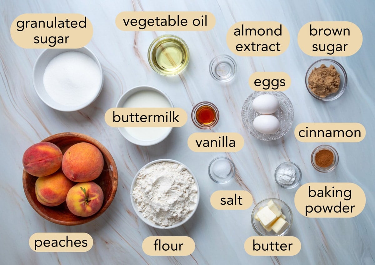 Overhead view of the ingredients for peach muffins arranged on a marble surface, including fresh peaches, flour, granulated sugar, brown sugar, buttermilk, vegetable oil, butter, eggs, vanilla extract, almond extract, baking powder, salt, and cinnamon, with each ingredient labeled.