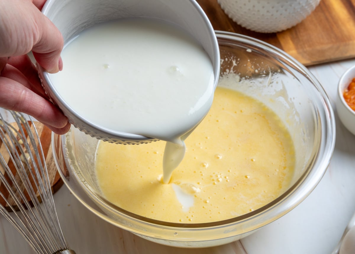 Pouring buttermilk into a bowl of whisked eggs, sugar, butter, and oil to make the wet batter for peach muffins, with a whisk, peach purée, and baking ingredients visible on the countertop.