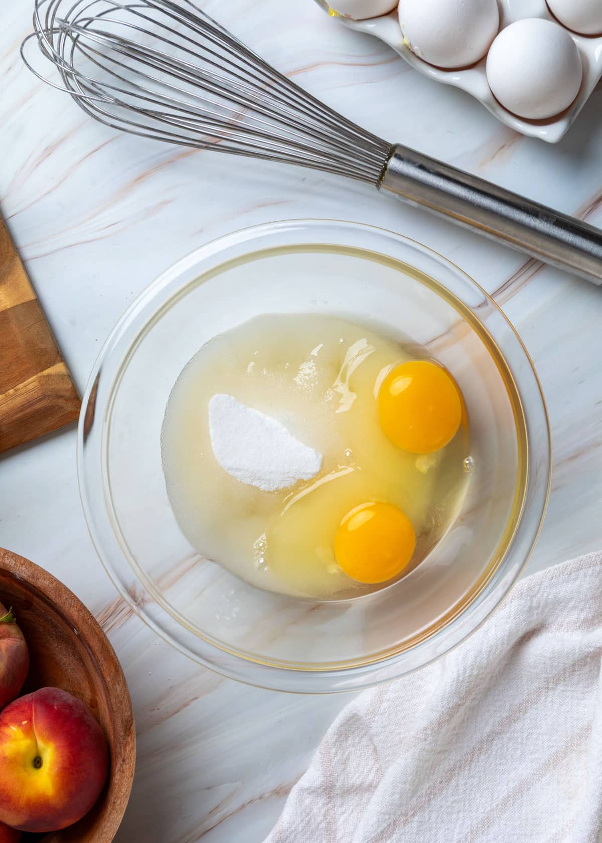 Overhead view of eggs, granulated sugar, and vegetable oil in a glass mixing bowl before whisking together for the peach muffin batter, with a whisk, whole peaches, and extra eggs nearby.