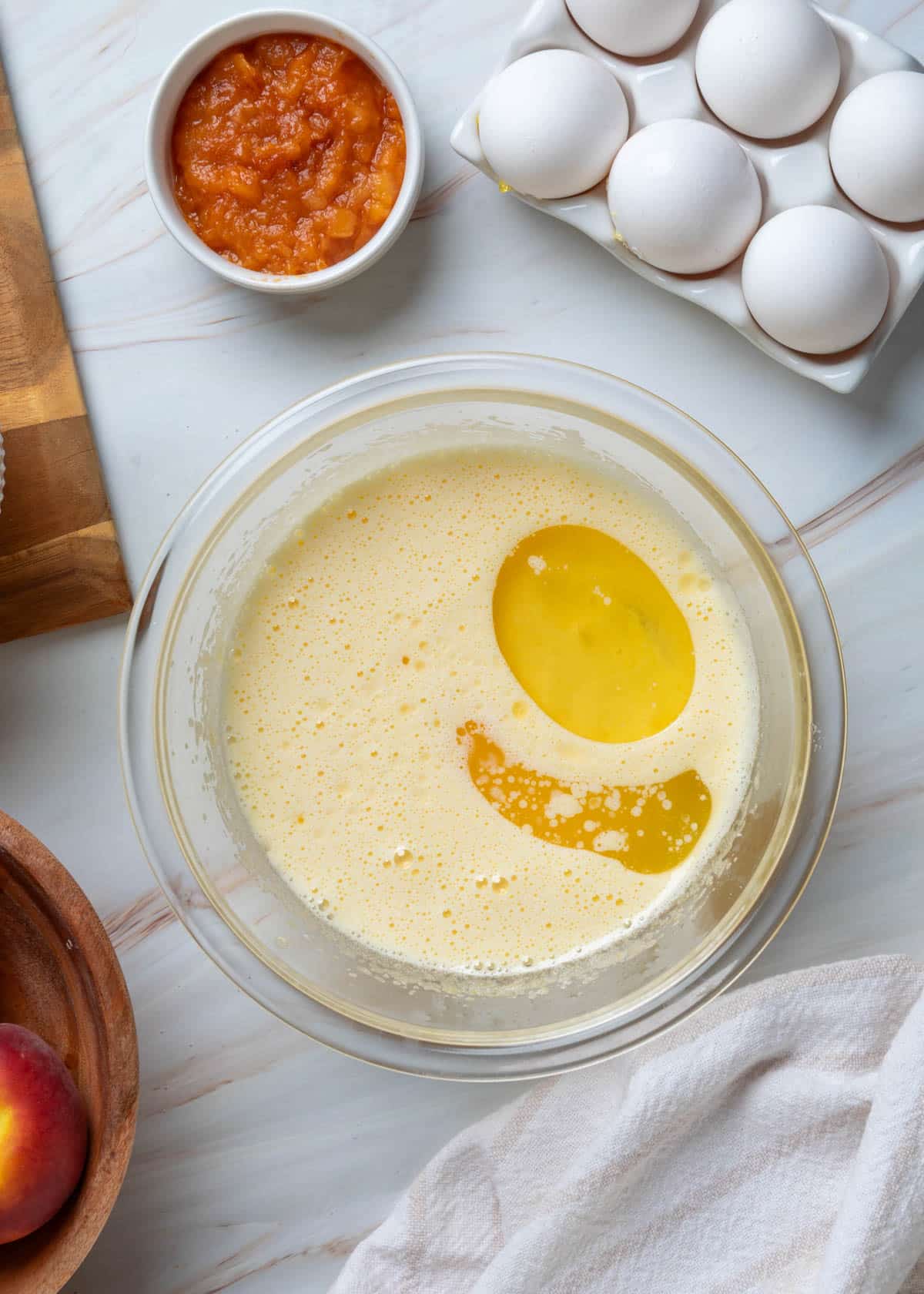 Overhead view of beaten eggs and sugar with melted butter and vegetable oil added to a glass mixing bowl, with peach purée and whole eggs nearby, ready to be whisked into the wet ingredients for peach muffins.