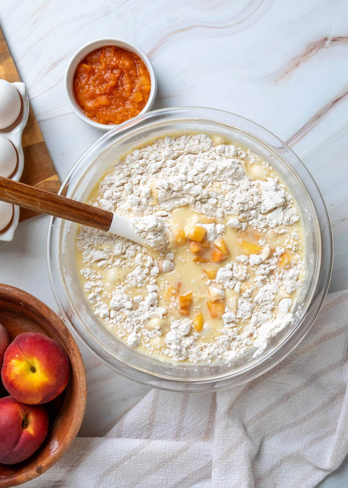 Dry ingredients and diced peaches added to the wet batter in a glass mixing bowl, ready to be folded together with a spatula to make peach muffin batter.