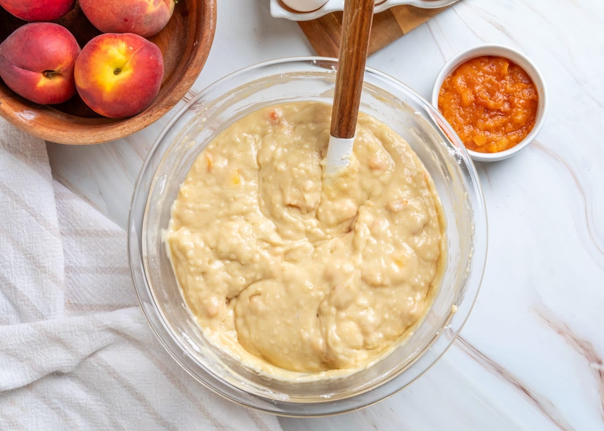 Overhead view of thick peach muffin batter after the wet and dry ingredients have been gently folded together, with visible pieces of fresh peach, a spatula, whole peaches, and peach purée nearby.