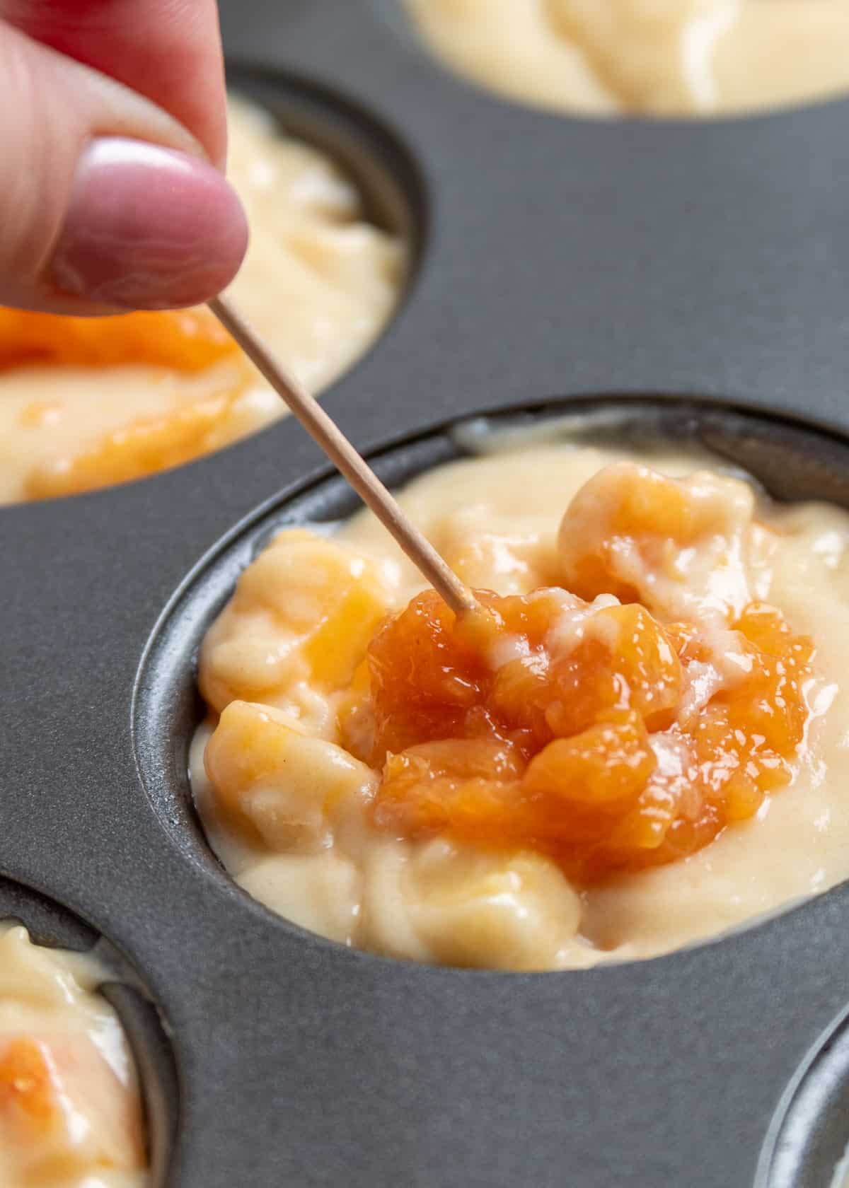 Using a toothpick to swirl a spoonful of peach compote into the top of unbaked peach muffin batter in a muffin pan before baking.