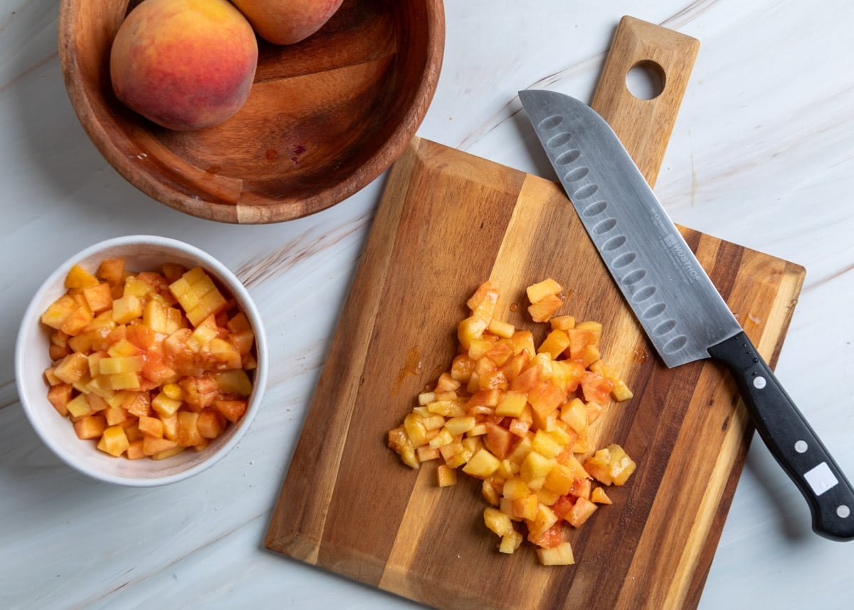 Overhead view of diced fresh peaches on a wooden cutting board beside a chef’s knife, with a bowl of diced peaches, whole peaches in a wooden bowl, and a marble work surface in the background.
