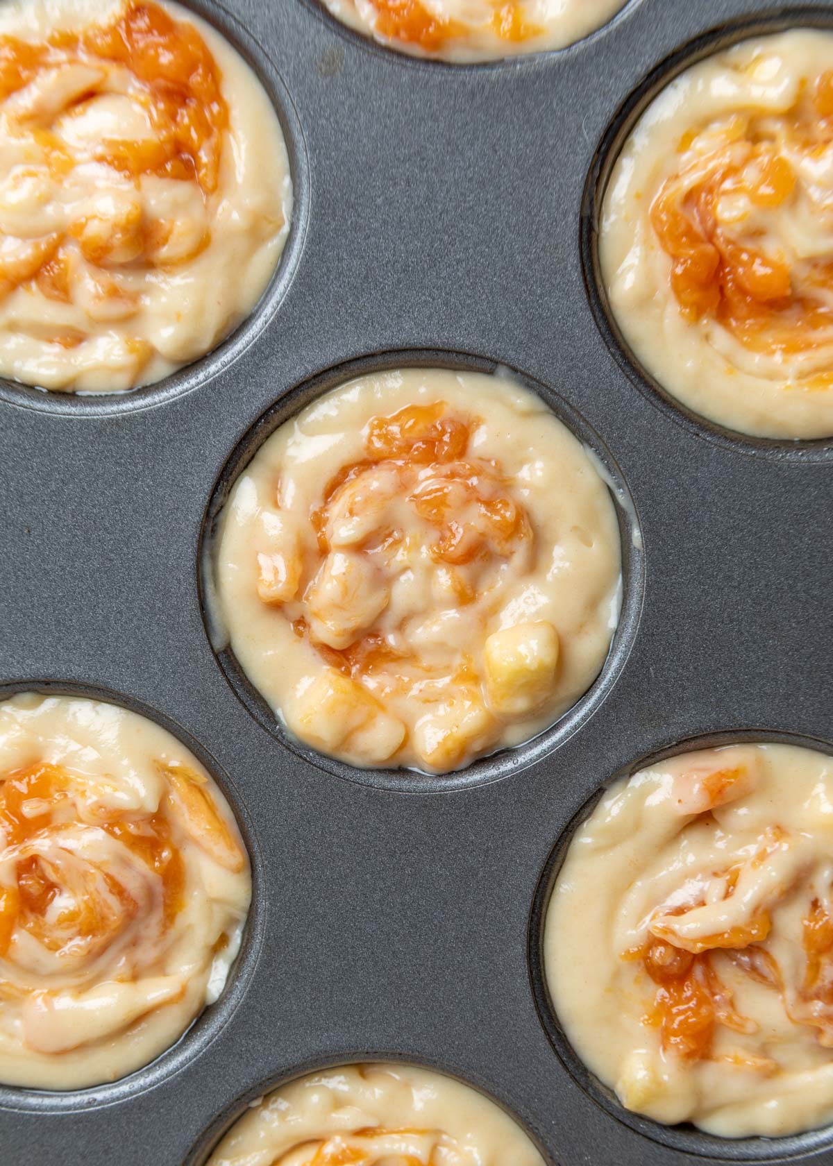 Overhead close-up of a muffin pan filled with unbaked peach muffin batter, each muffin topped with a swirled layer of peach compote before baking.