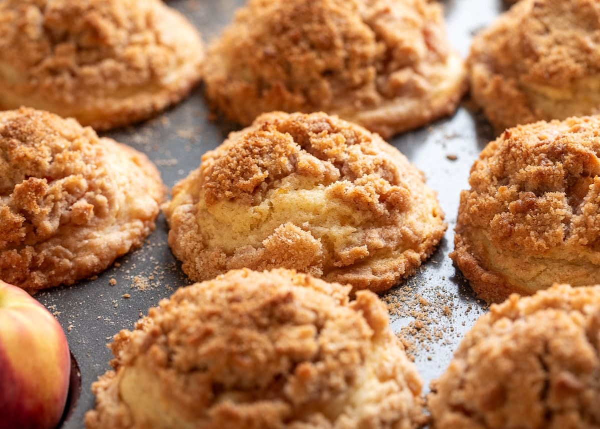 Freshly baked peach muffins with tall, domed tops and a buttery cinnamon streusel topping cooling in the muffin pan, with crisp golden edges and visible crumb scattered around the muffins.