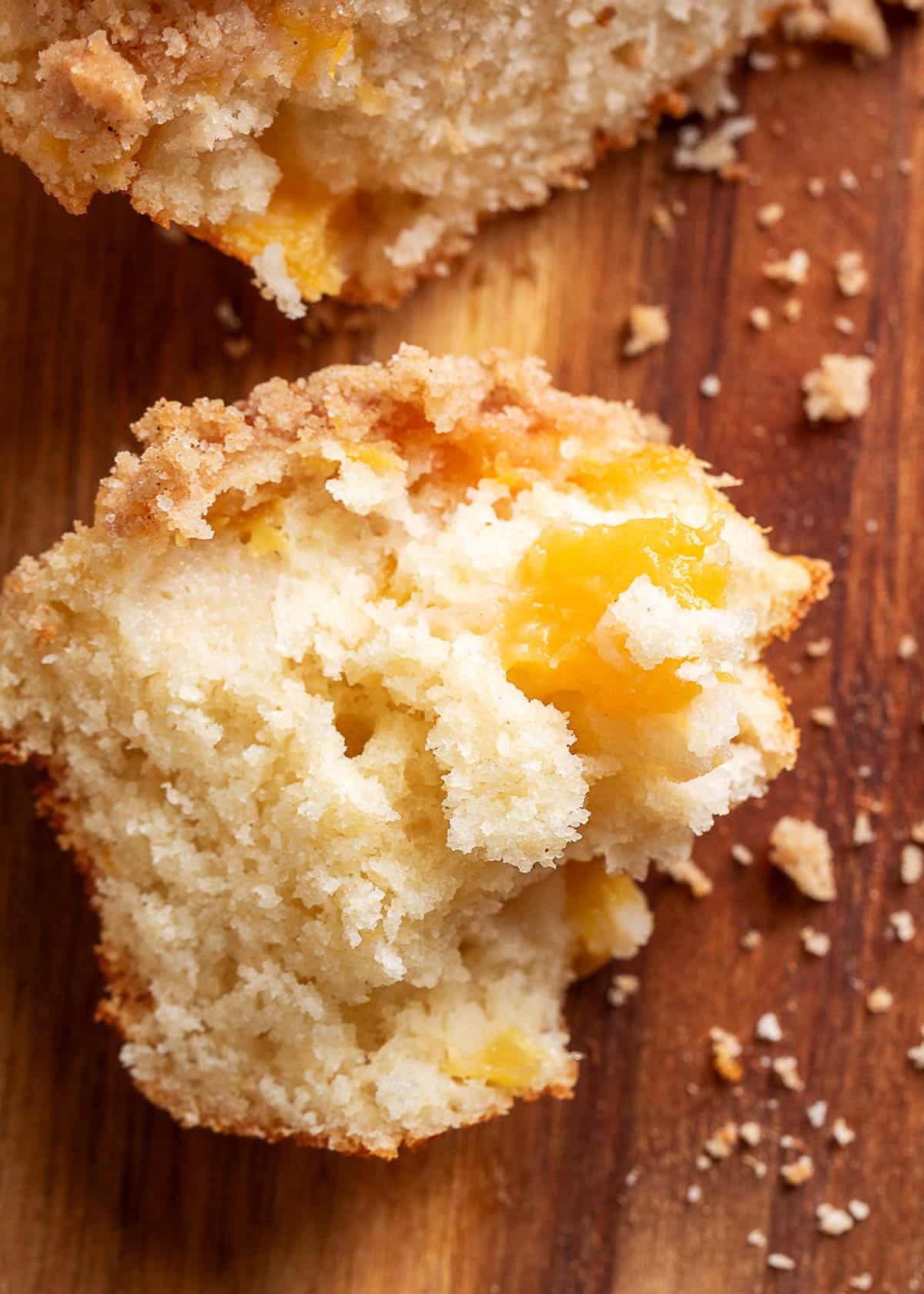 A peach streusel muffin broken open to reveal a soft, fluffy crumb with juicy chunks of fresh peach throughout, topped with a buttery cinnamon streusel and surrounded by scattered crumbs on a wooden cutting board.