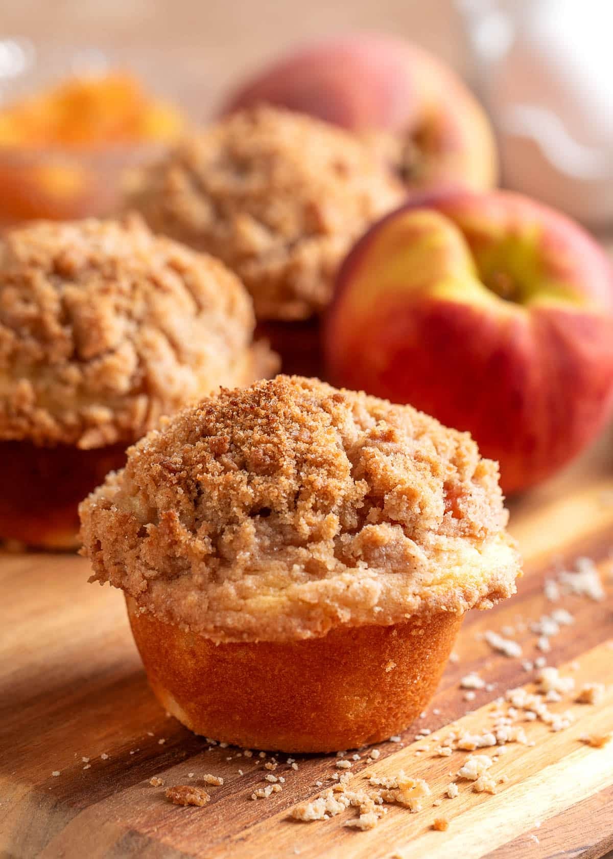 A peach streusel muffin topped with a thick layer of buttery cinnamon crumb topping sits on a wooden board, with fresh peaches, additional muffins, and scattered streusel crumbs softly blurred in the background.