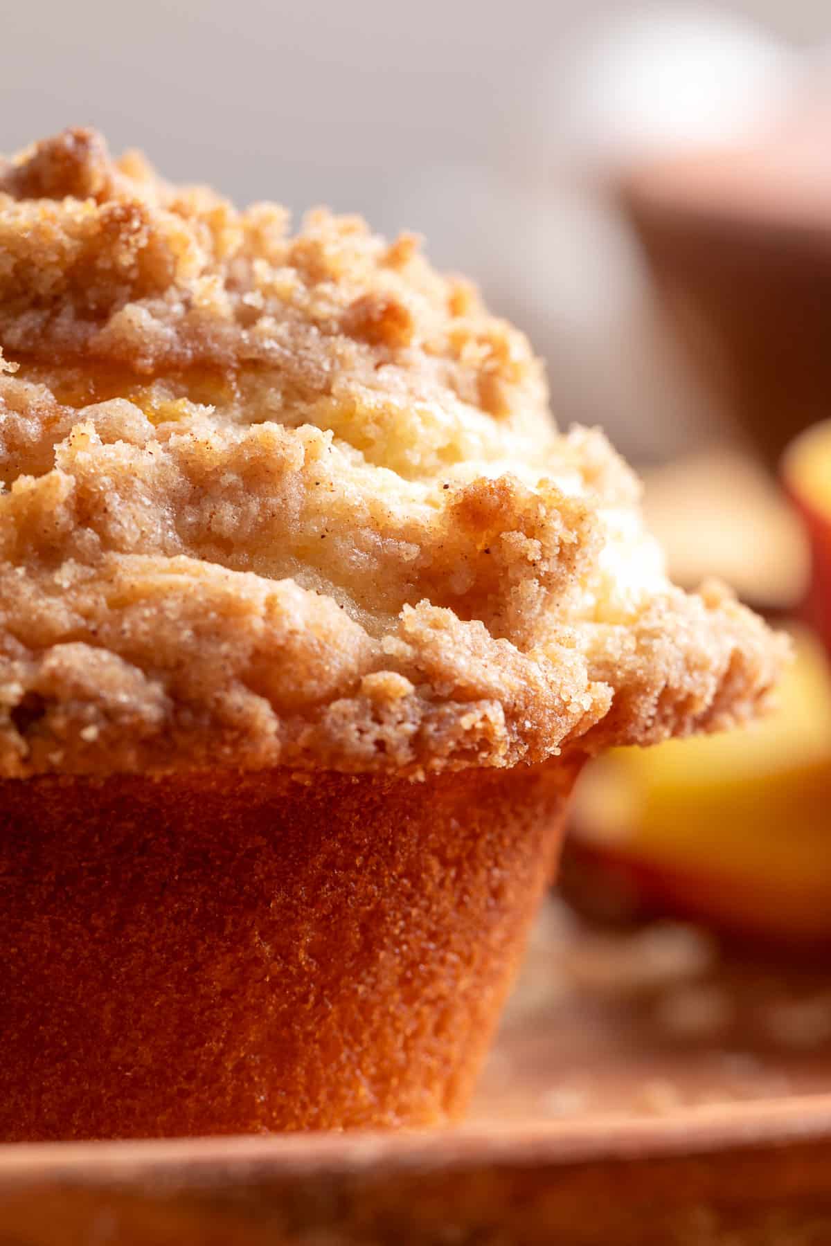 A close-up of a peach streusel muffin with a golden-brown, buttery crumb topping and tender cake, highlighting the crisp streusel texture with fresh peaches softly blurred in the background.