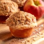 A peach streusel muffin with a tall, buttery cinnamon crumb topping sits on a wooden board with scattered streusel crumbs, while fresh peaches and additional muffins are softly blurred in the background.