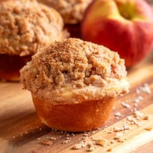 A peach streusel muffin with a tall, buttery cinnamon crumb topping sits on a wooden board with scattered streusel crumbs, while fresh peaches and additional muffins are softly blurred in the background.