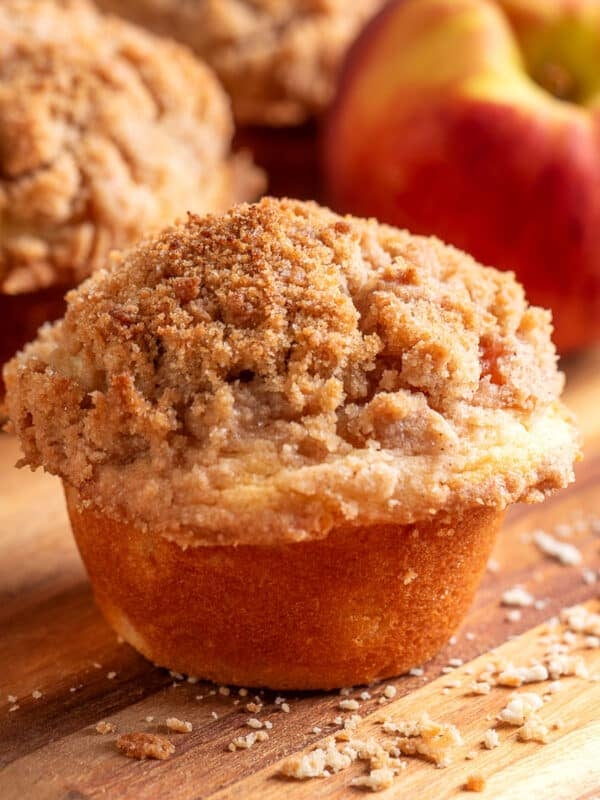 A peach streusel muffin with a tall, buttery cinnamon crumb topping sits on a wooden board with scattered streusel crumbs, while fresh peaches and additional muffins are softly blurred in the background.