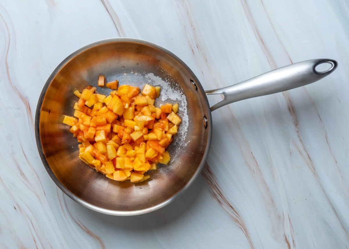 Overhead view of diced peaches and sugar in a stainless steel saucepan, ready to be cooked into a peach filling on a light marble work surface.