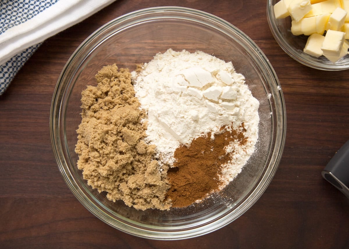 Overhead view of streusel ingredients in a glass bowl, including flour, brown sugar, cinnamon, and cold butter cubes before cutting the butter into the dry ingredients.