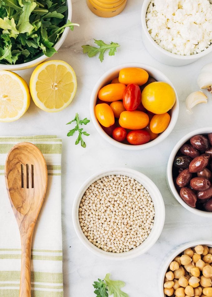 overhead photo of bowls of pearled couscous, cherry tomatoes, feta, olives, and chickpeas