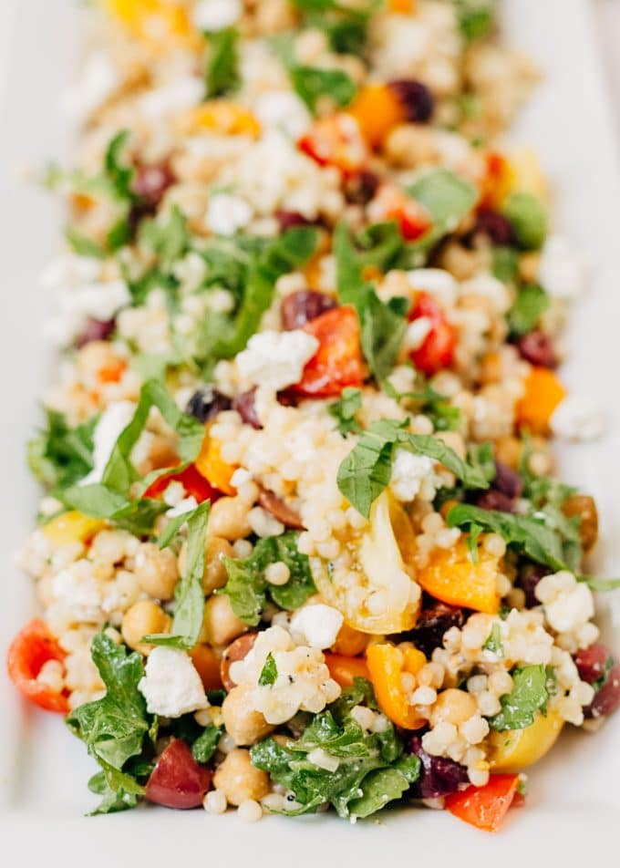 side view of israeli couscous salad with arugula, tomatoes, and feta on a white platter