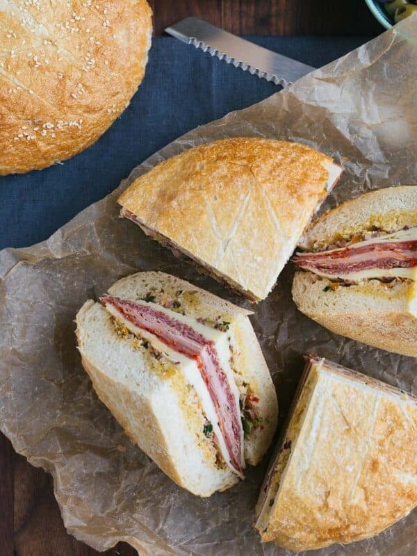 Overhead view of a round muffuletta sandwich cut into quarters, revealing layered meats, cheeses, and olive salad on parchment paper, with a whole loaf of sesame bread and a bowl of olives nearby.