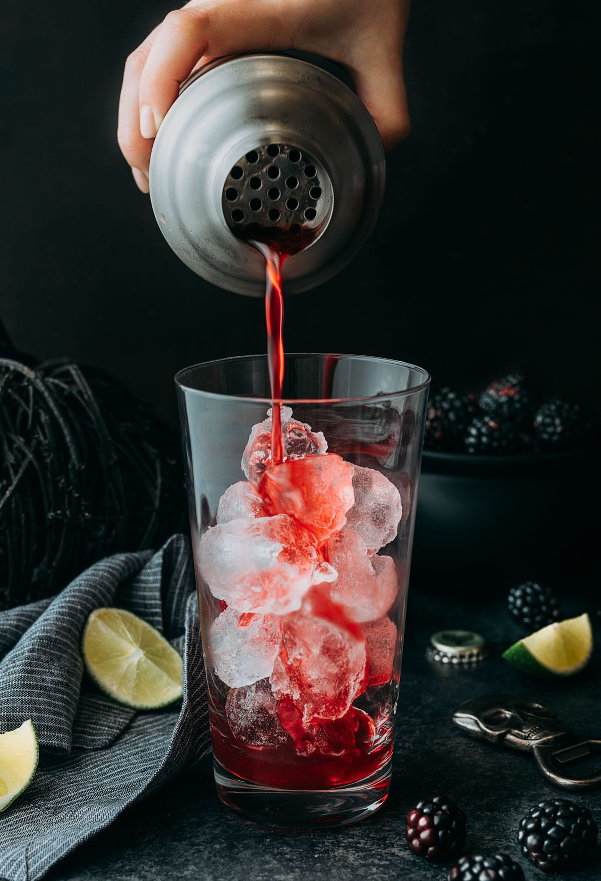 straining a blackberry el diablo cocktail into an ice-filled highball glass from a cocktail shaker