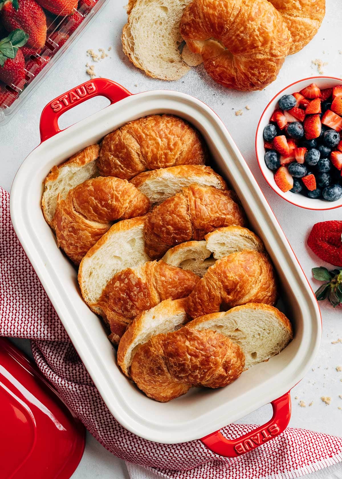 overhead photo of sliced croissants in a rectangular baking dish, next to a bowl of blueberries and chopped strawberries