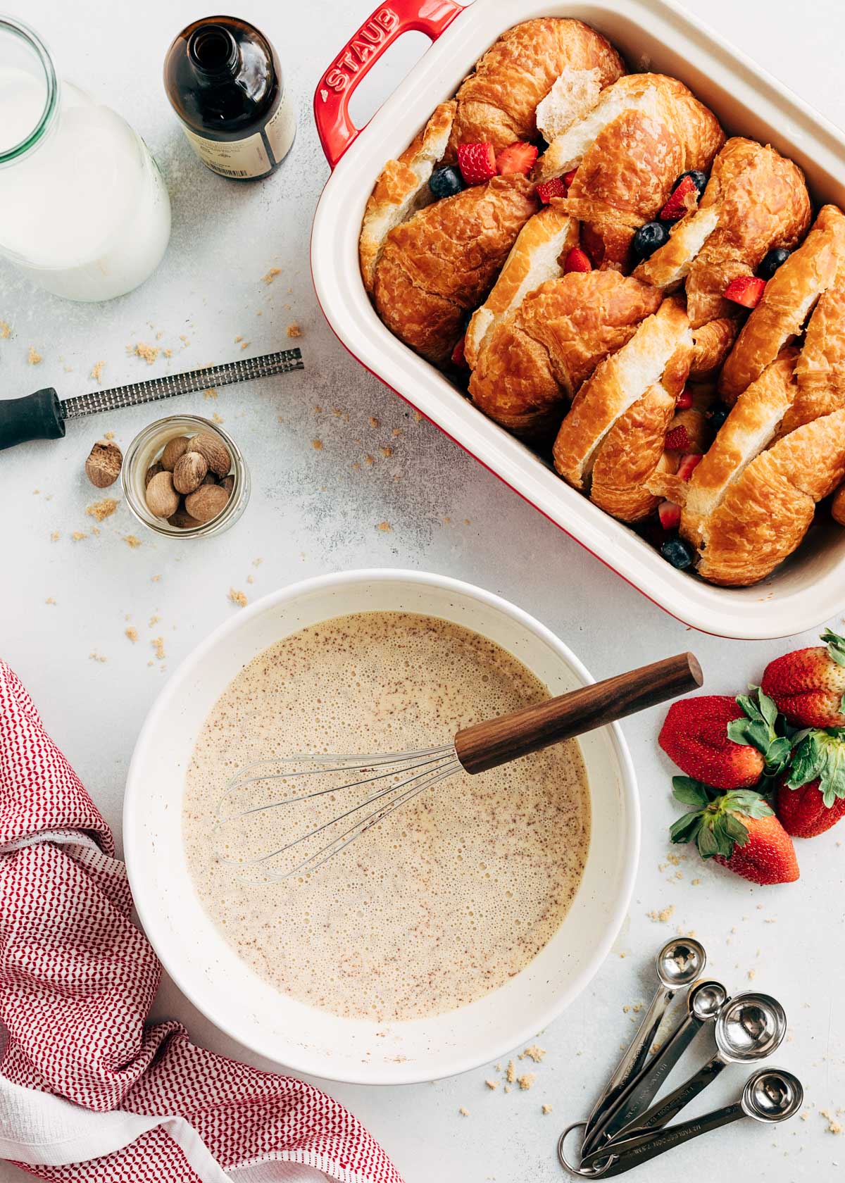 bowl with a whisk containing French Toast custard and a baking dish holding sliced croissants and berries