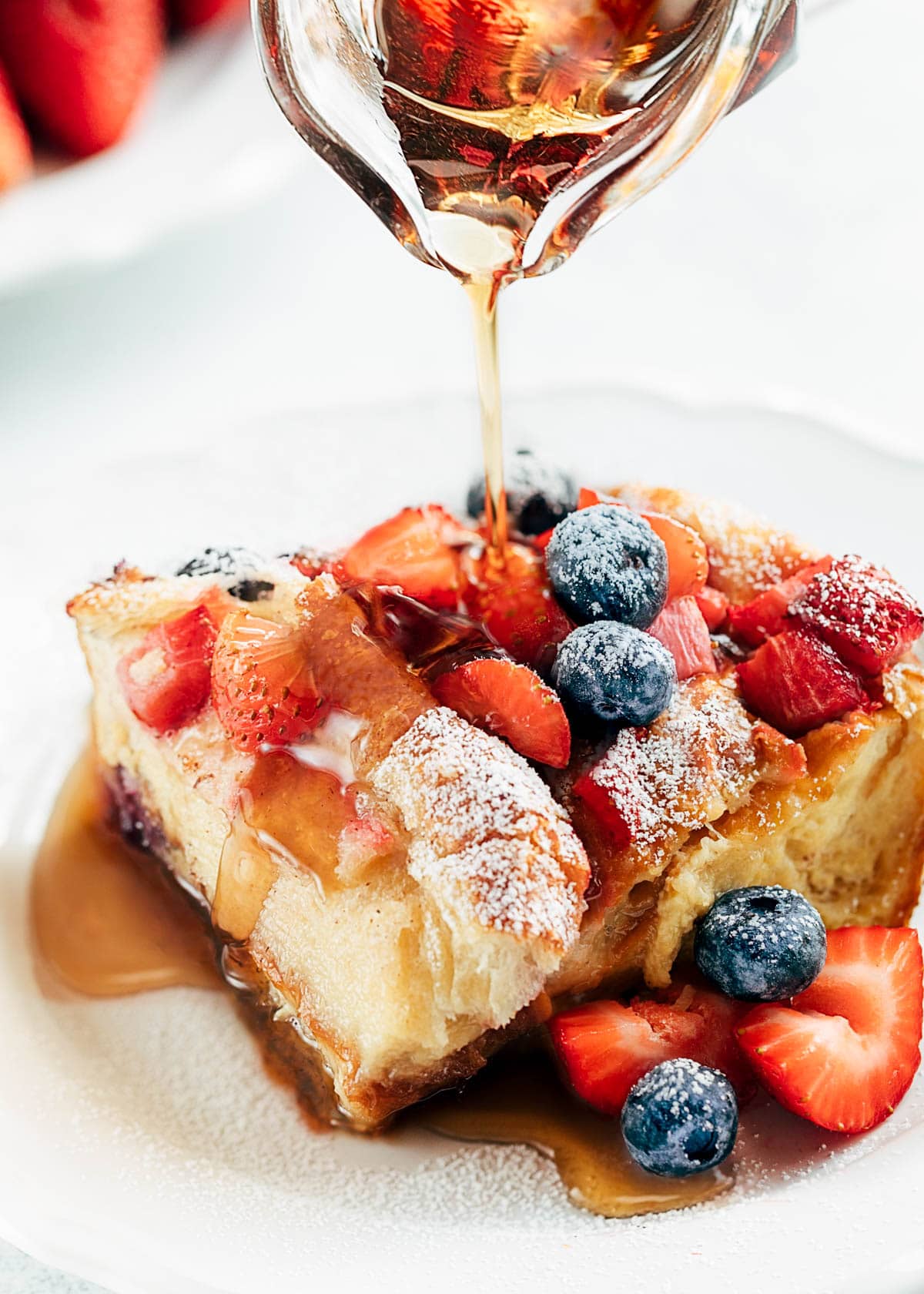 wedge of croissant french toast bake with berries on a white plate, with rum maple syrup being poured over the top