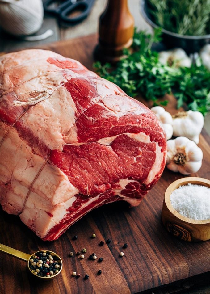 uncooked prime rib on a board with garlic, herbs, and a bowl of sea salt