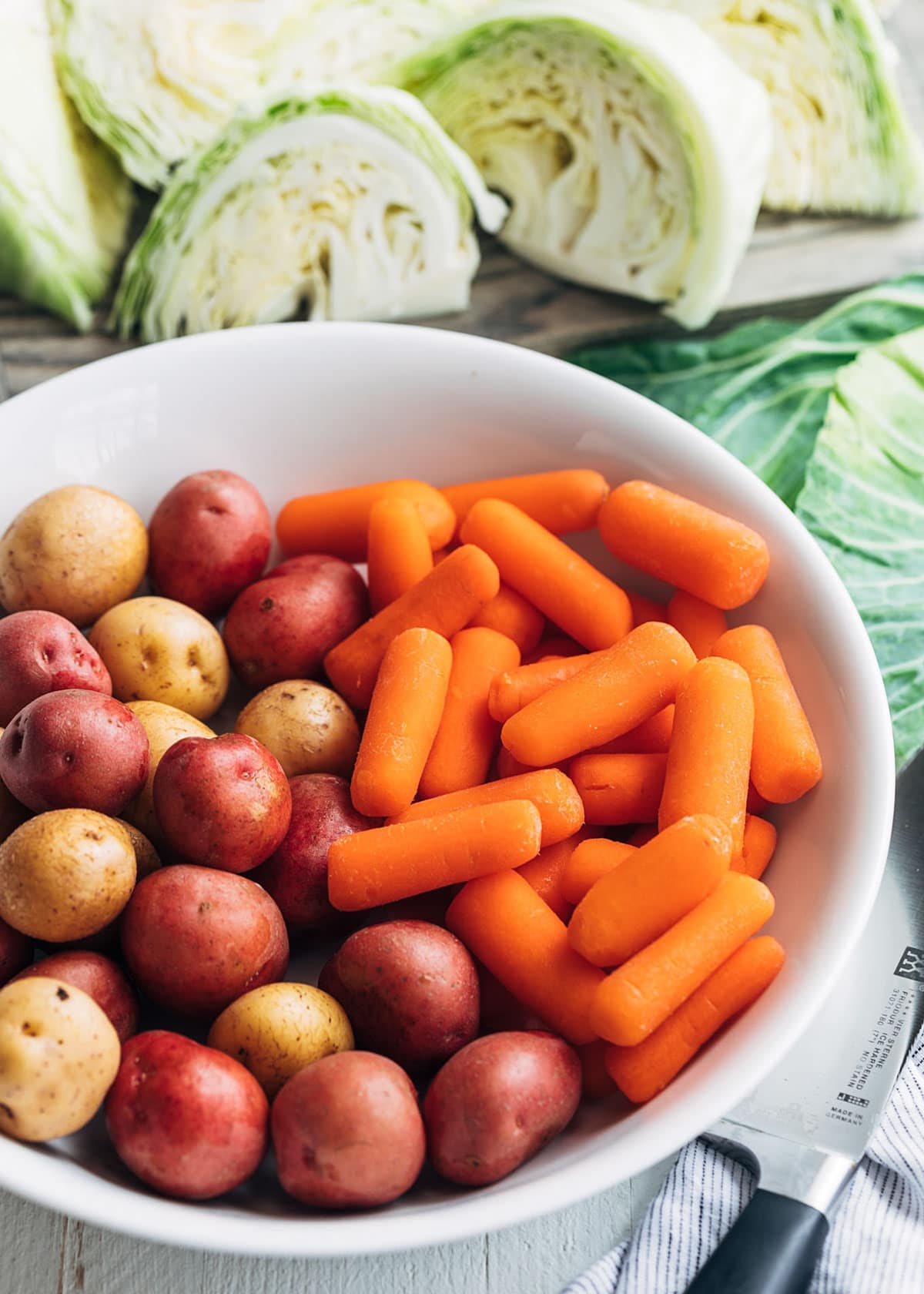 small potatoes and baby carrots in a white bowl surrounded by green cabbage wedges