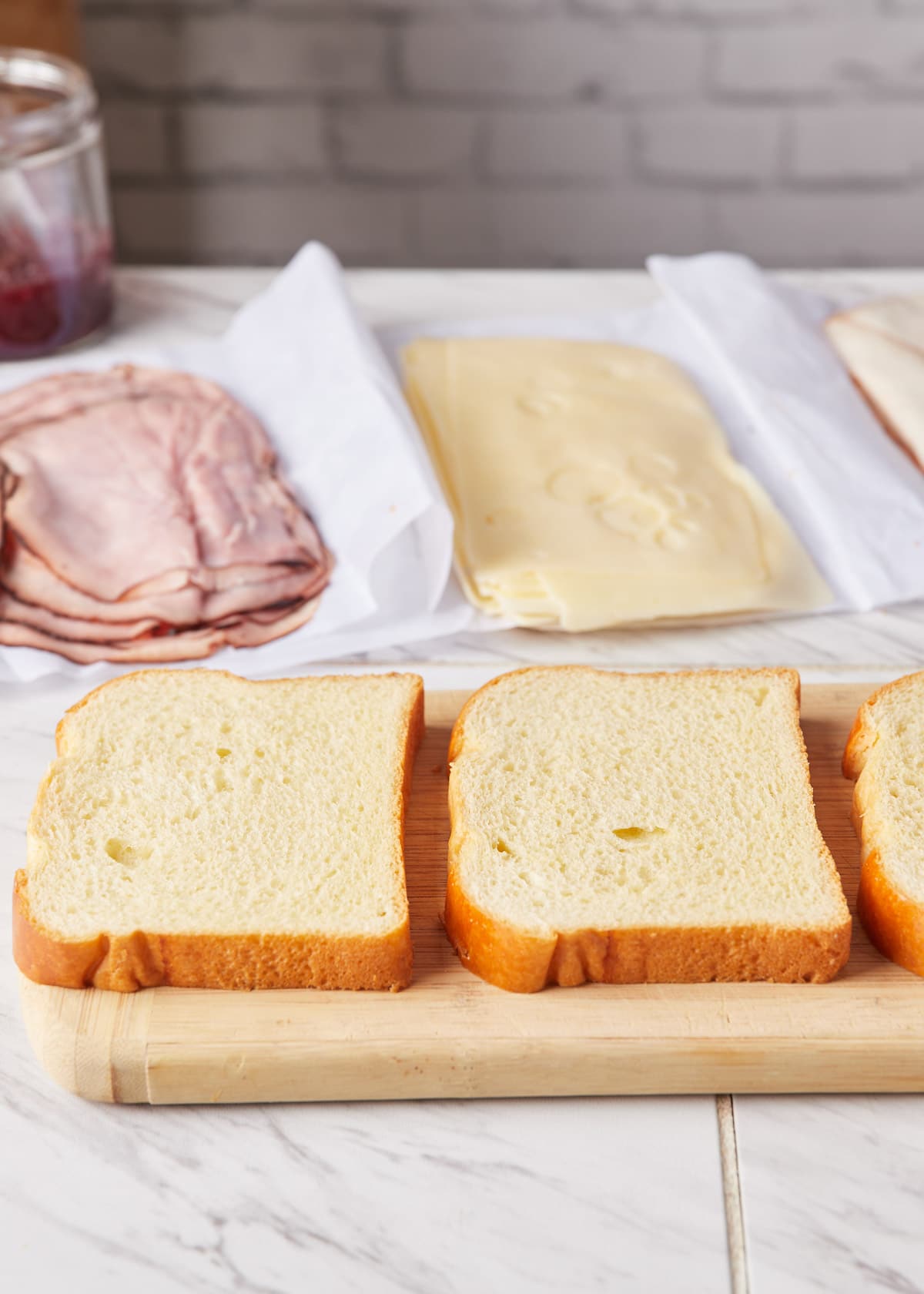 Slices of white bread on a wooden cutting board, in front of sliced ham, swiss, and turkey on deli paper