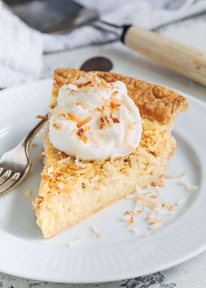 slice of coconut custard pie on a plate with whipped cream and toasted coconut flakes