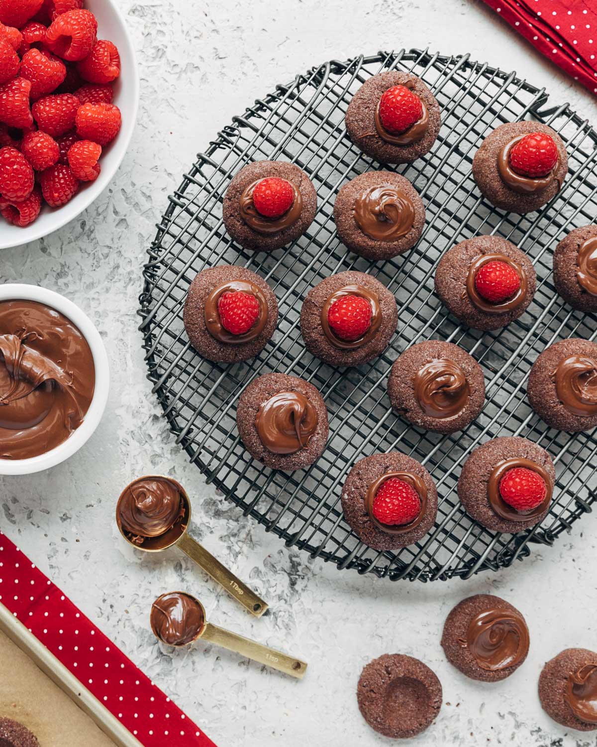 filled chocolate hazelnut thumbprint cookies on a cooling rack with bowls of raspberries and chocolate hazelnut spread