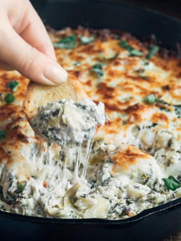 dipping a crostini toast into a cast iron pan of spinach artichoke dip, with a cheese pull