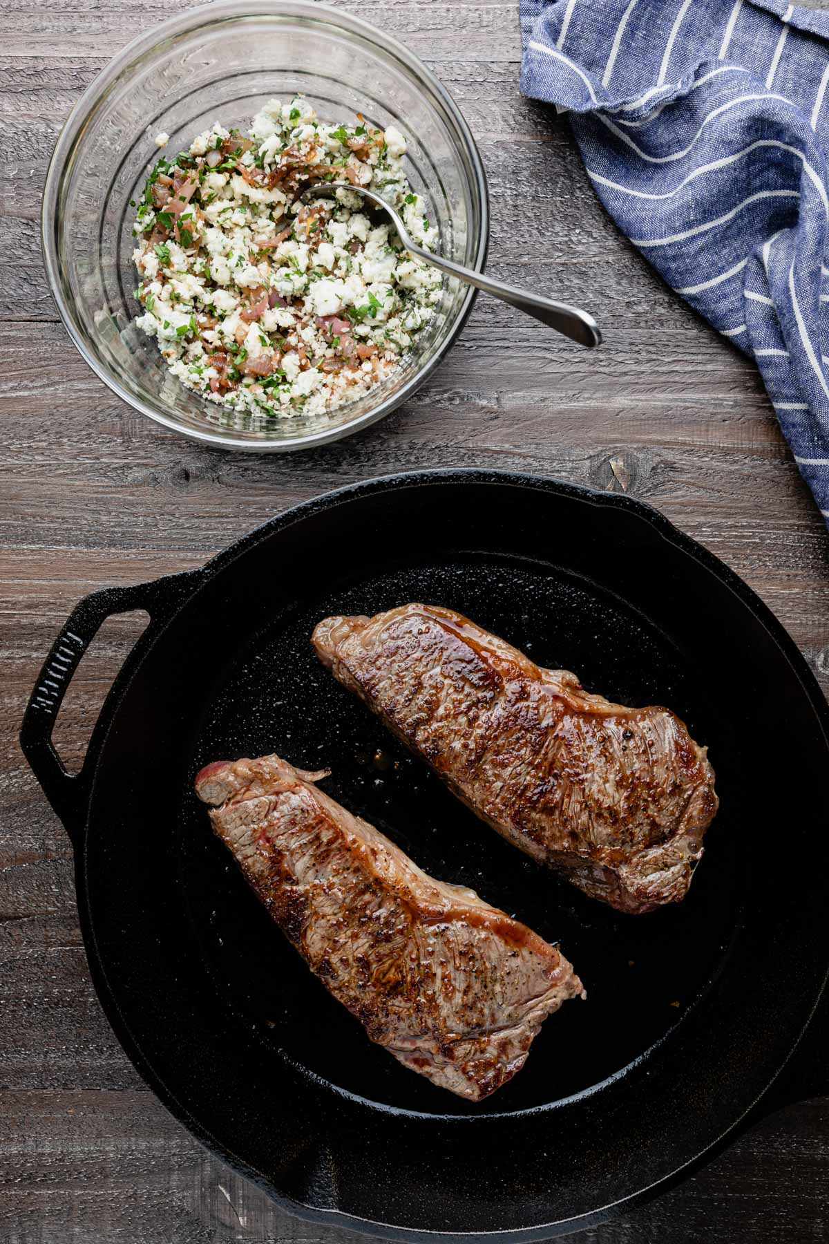 seared strip steaks in a cast iron pan next to a bowl of blue cheese crust mixture