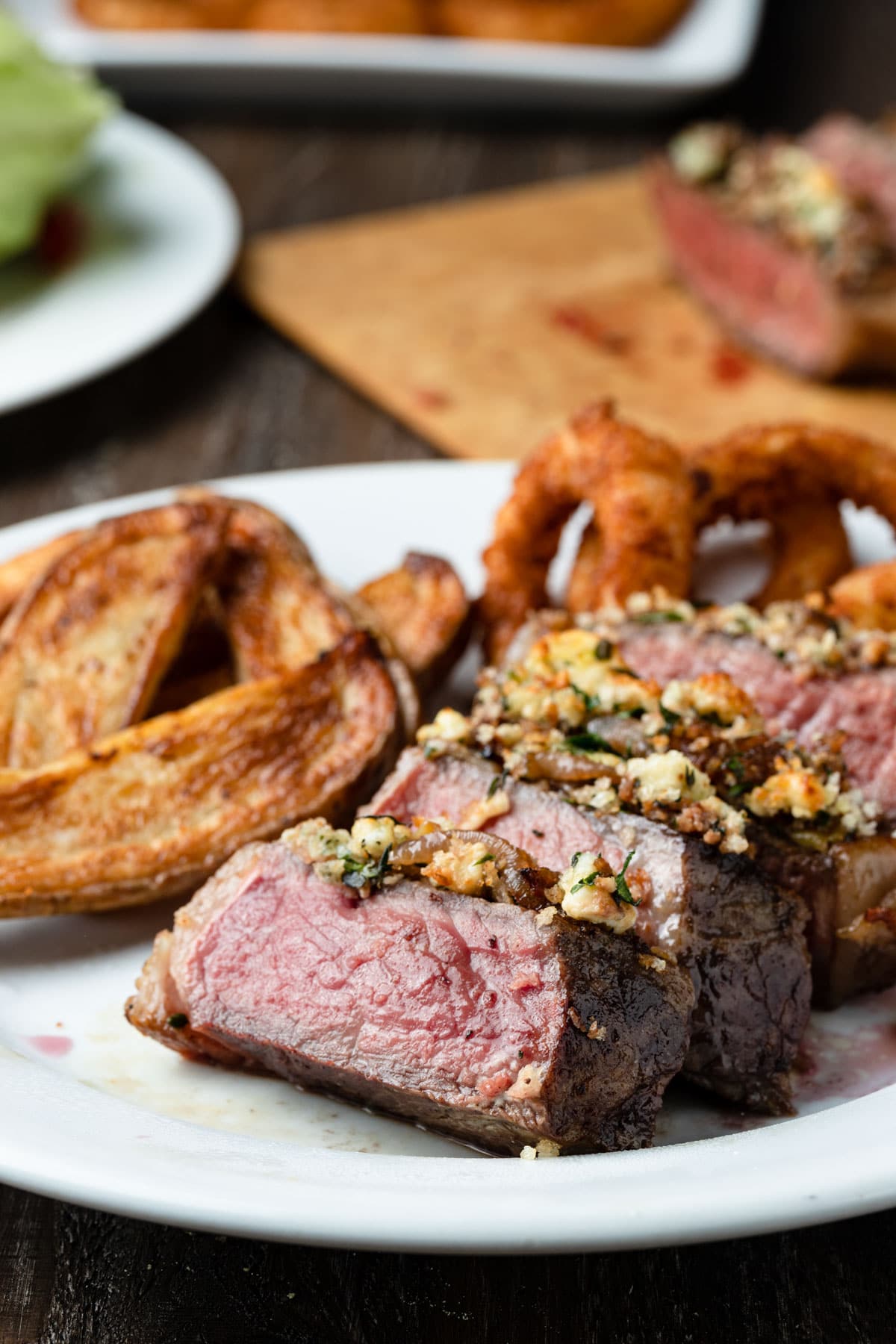 blue cheese crusted new york strip steak on a white plate with french fries and onion rings