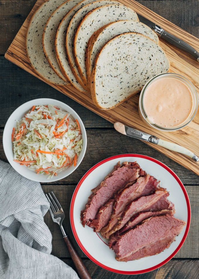 sliced corned beef on a plate next to a bowl of coleslaw and loaf of seeded rye bread