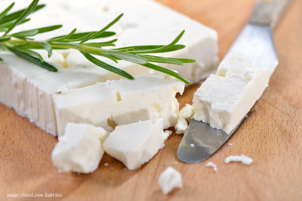 feta block on a wood board, partially crumbled with a cheese knife and sprig of rosemary