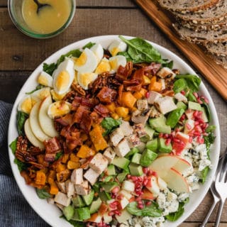 overhead of a fall harvest cobb salad in a white bowl next to a jar of maple dijon vinaigrette and a board of whole grain bread