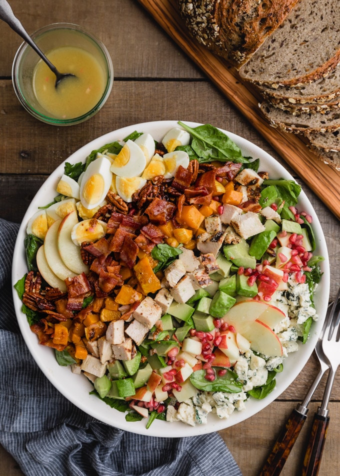 overhead of a fall harvest cobb salad in a white bowl next to a jar of maple dijon vinaigrette and a board of whole grain bread