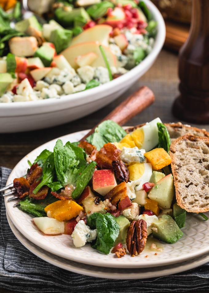 side view of a Cobb salad with apples, pecans, and butternut squash on an ivory plate with whole grain bread