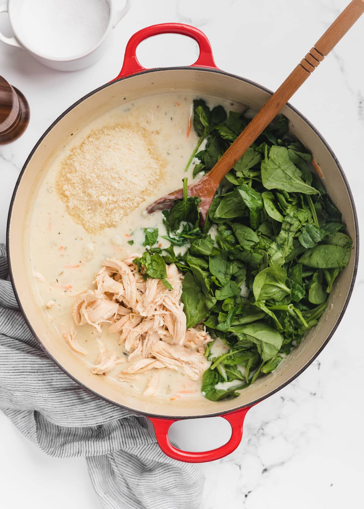 shredded rotisserie chicken and raw baby spinach are added to a Dutch oven of soup. A wooden spoon rests in the pot.