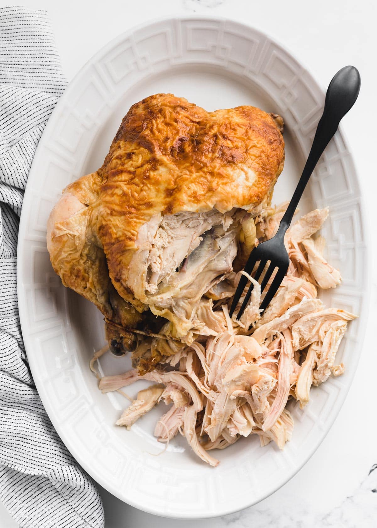 partially shredded rotisserie chicken on a white platter with a black fork
