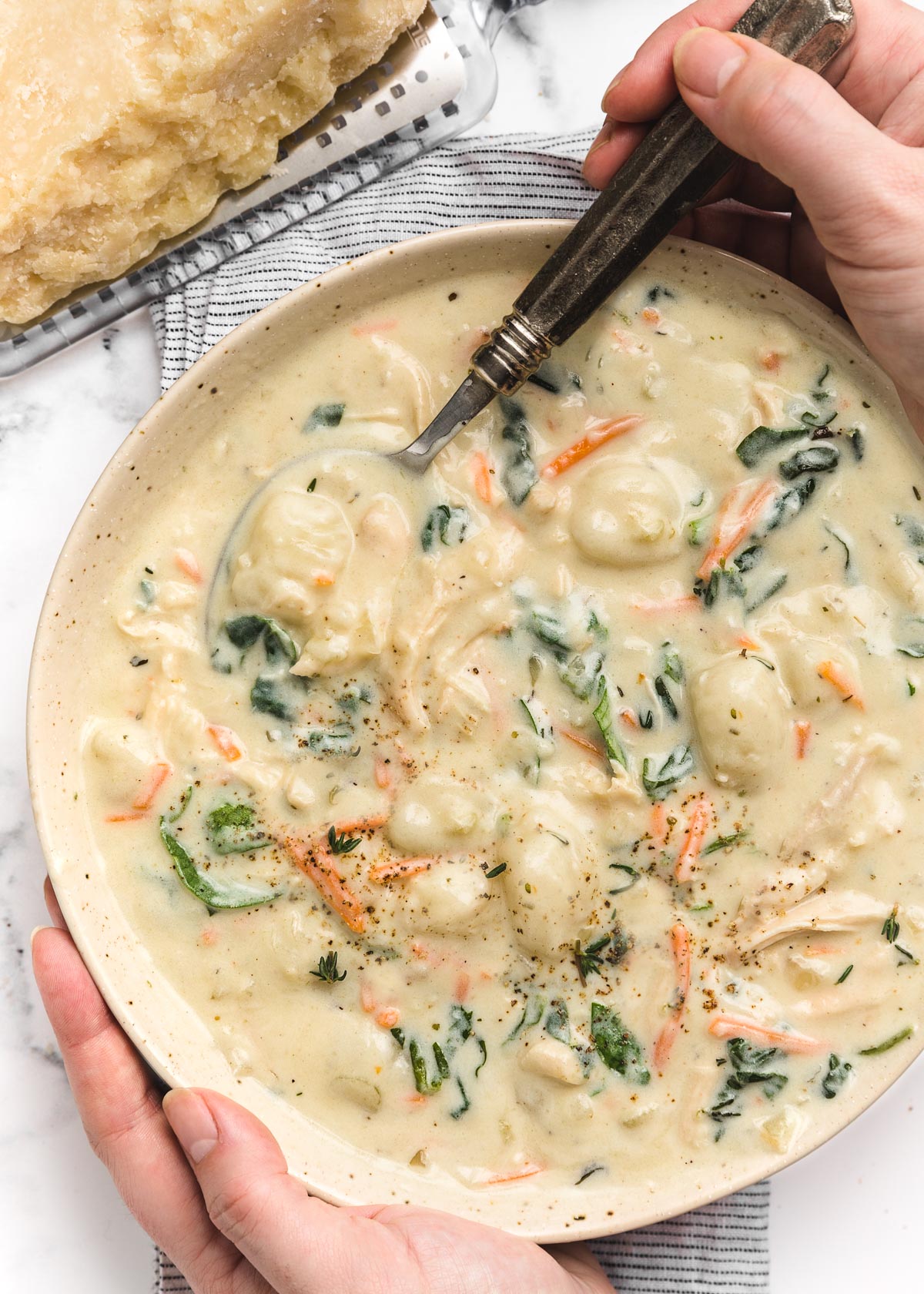 overhead photo of two hands holding a bowl of creamy chicken gnocchi soup. One hand holds a silver spoon.