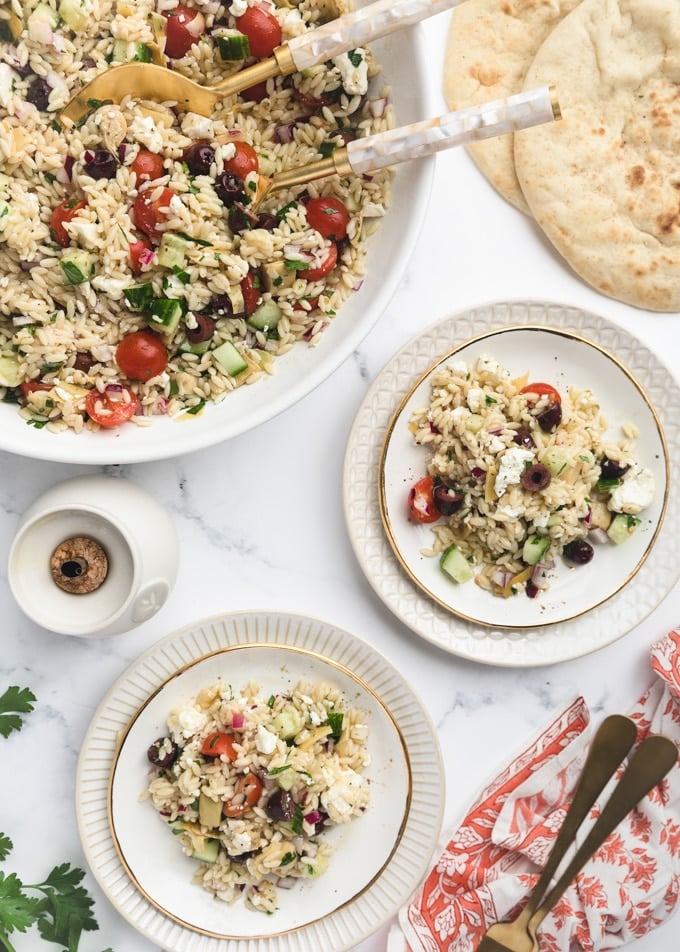 overhead of orzo salad served on white plates, next to a big bowl of the salad and pita bread rounds 